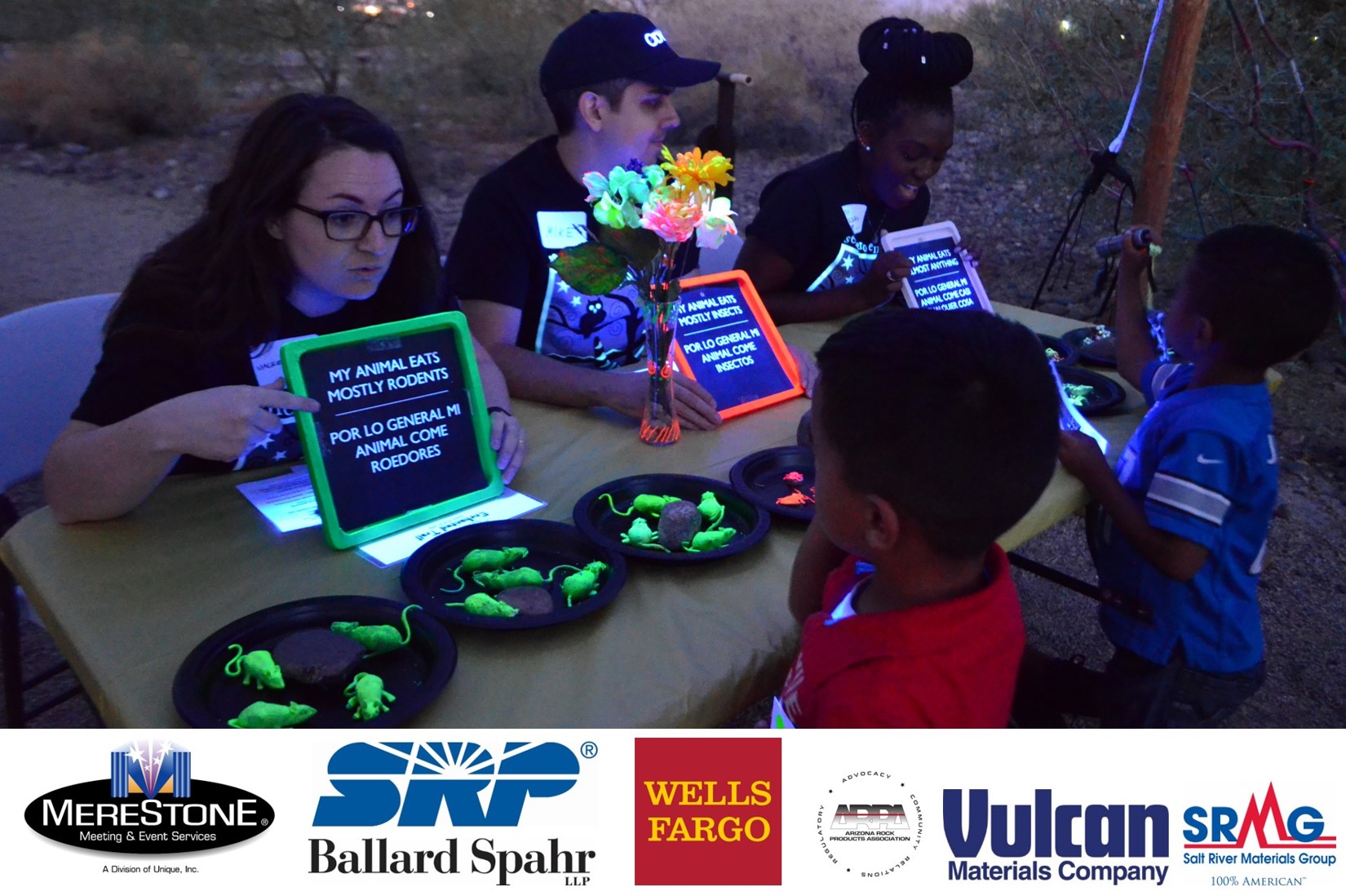 Volunteers point at signs about animal diets to children during the Enchanted Trail/Sendero Encantado event. Sponsor logos include Merestone, SRP, Ballard Spahr, Wells Fargo, Arizona Rock Products Association, Vulcan Materials Company, and Salt River Materials Group.