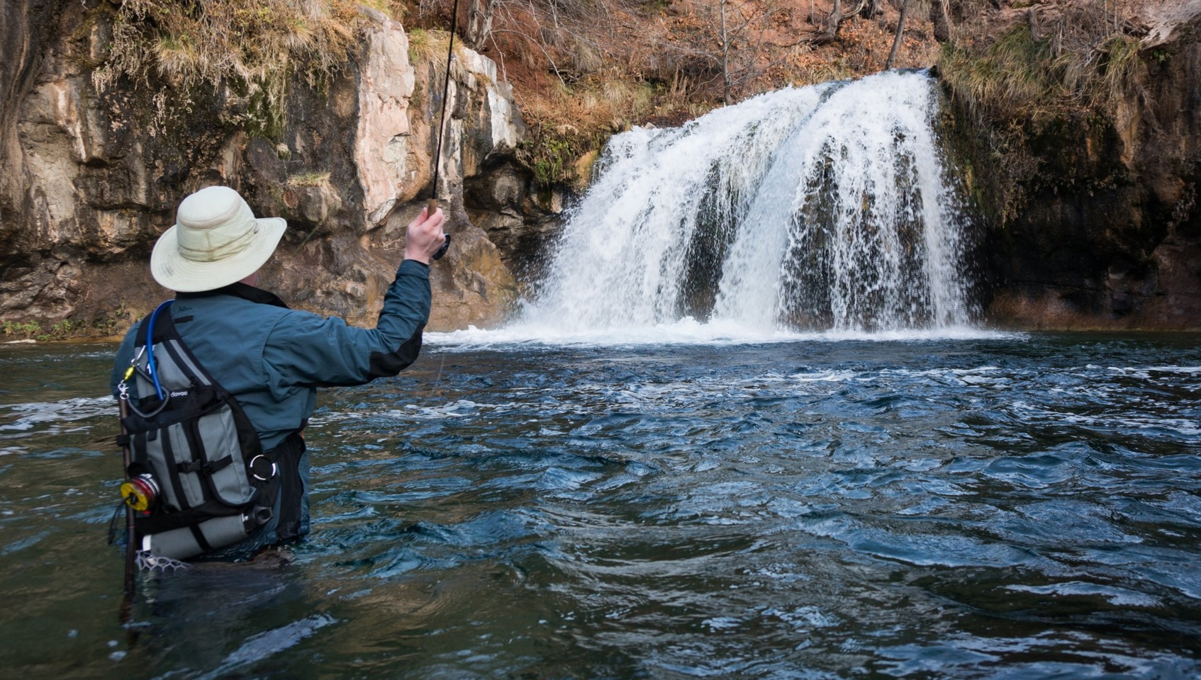 A man is standing in Fossil Creek casting his fishing line out towards a short waterfall.