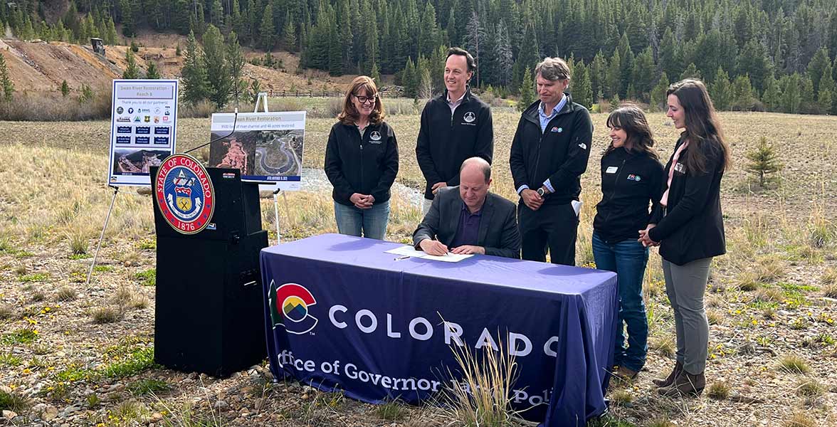 Five individuals stand behind the Governor Polis, who is seated at a table, as he signs a document.
