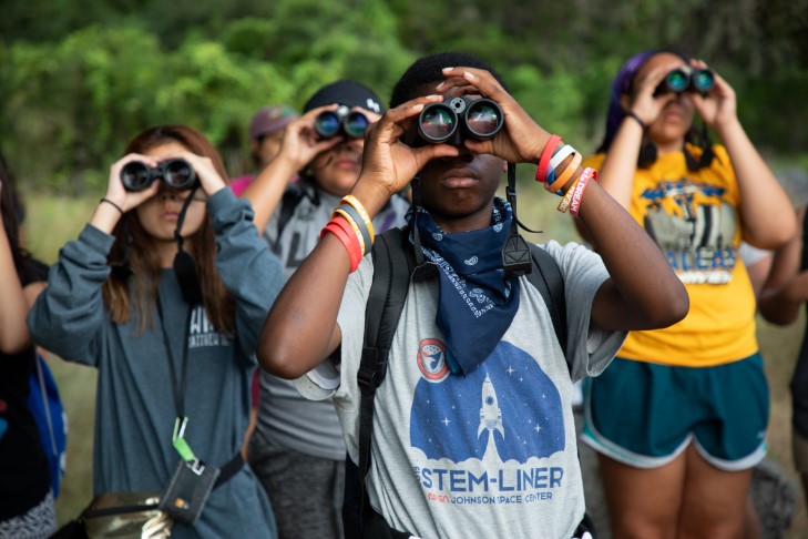 A group of young adults look past the camera with binoculars. The one in front is a black male, and behind him are other birders of color.