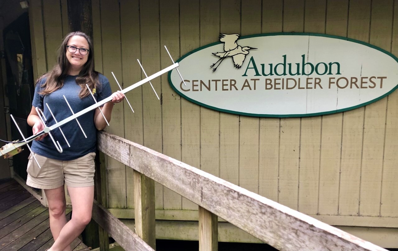 A woman with brown hair, blue shirt, beige shorts stands in front of the Beidler Forest entrance sign. She is holding a long metal rod with smaller metal pieces attached in a T-like formation. 