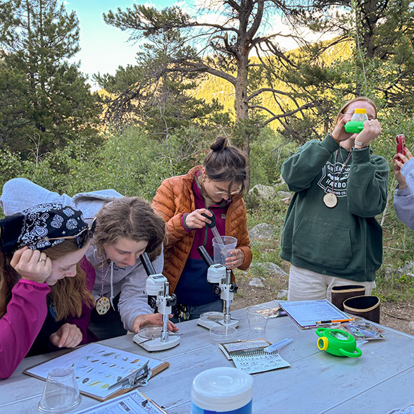 Five adults perform various activities around an outdoor table of science education materials.
