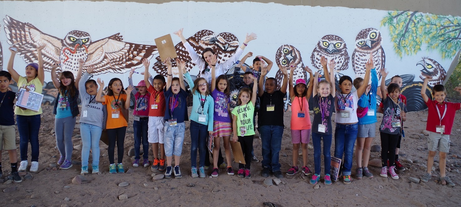 Students stand smiling in front of a Burrowing Owl mural.