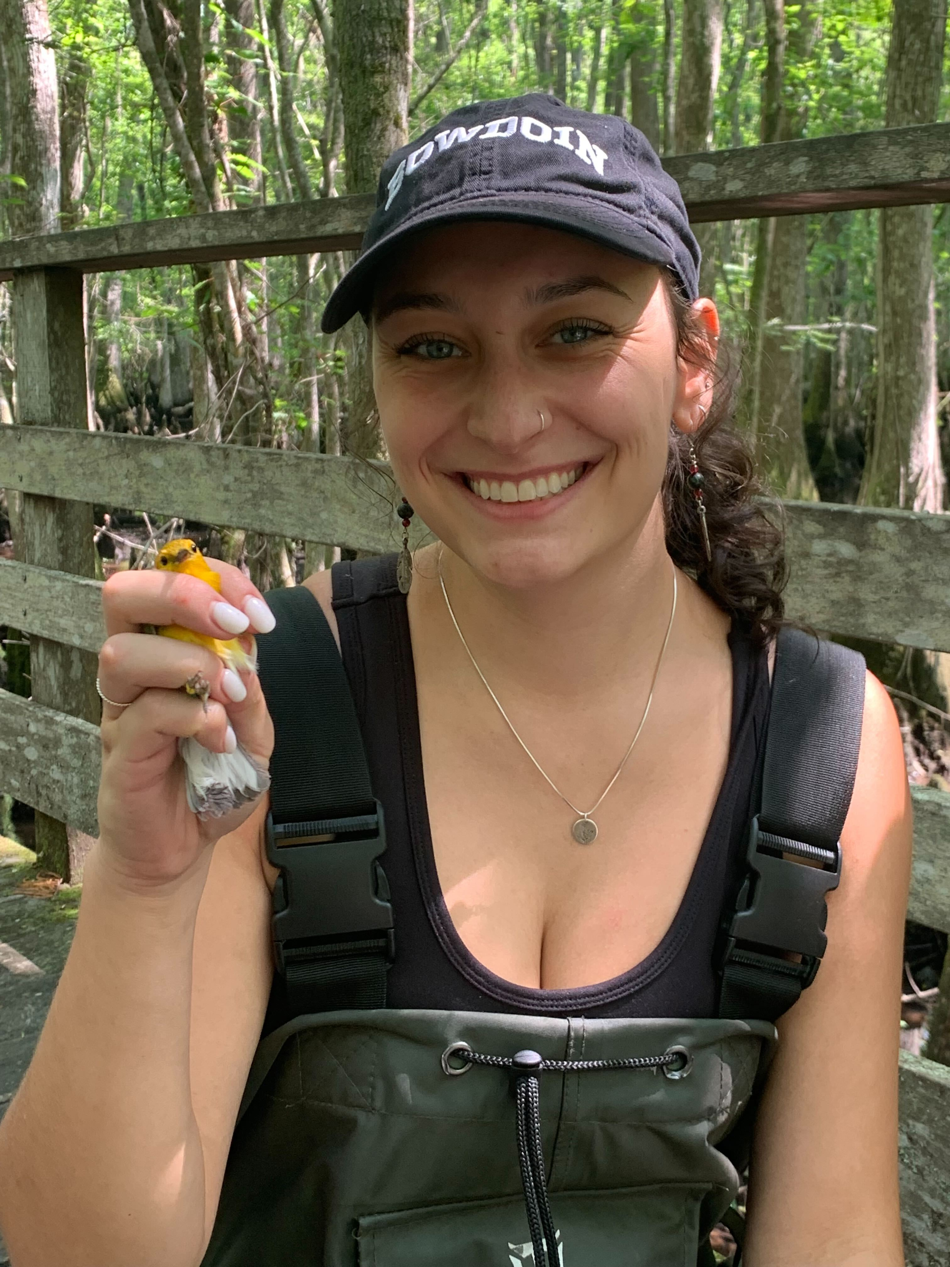 A woman with a blue-gray ball cap, brown hair, and dangling earrings is holding a small banded yellow bird in her right hand. 