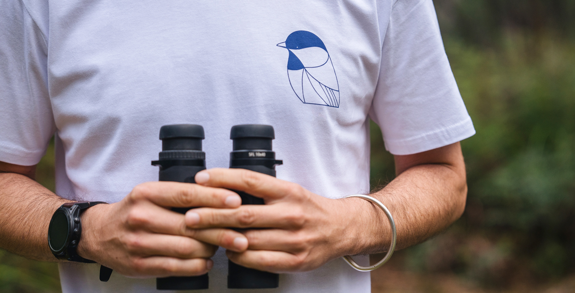 A person wears an Eggpicnic x The Birdsong Project shirt while holding a pair of binoculars.