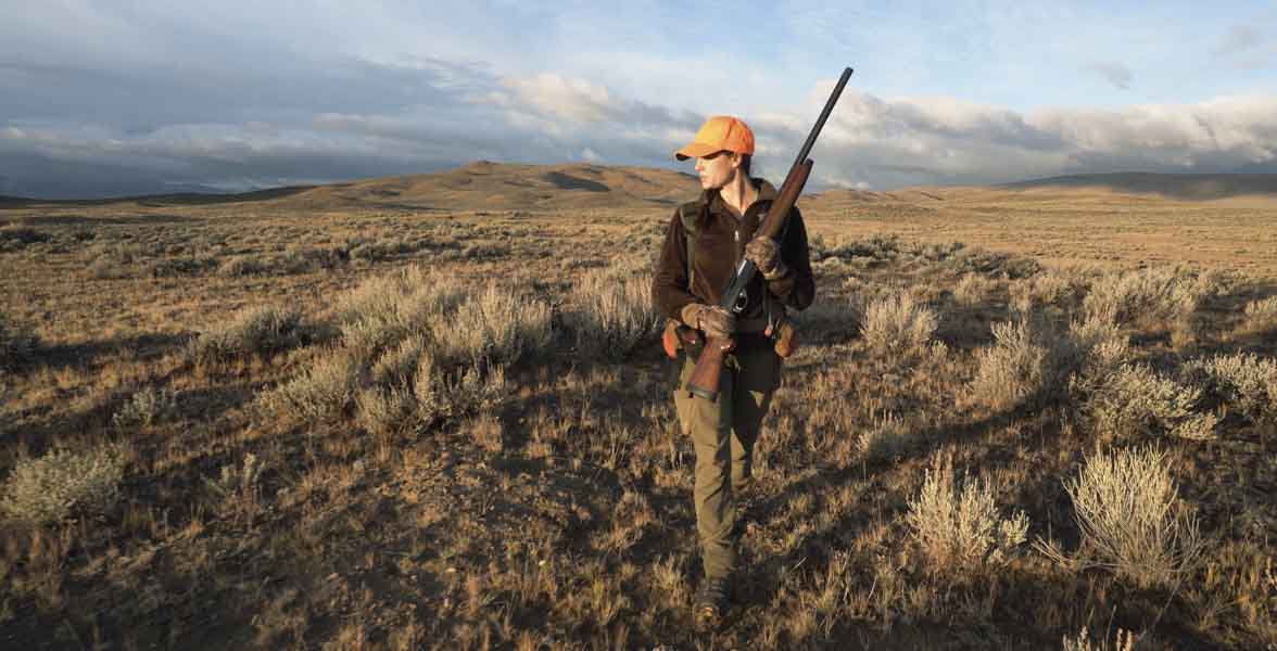A hunter walks in the sagebrush steppe with a rifle.