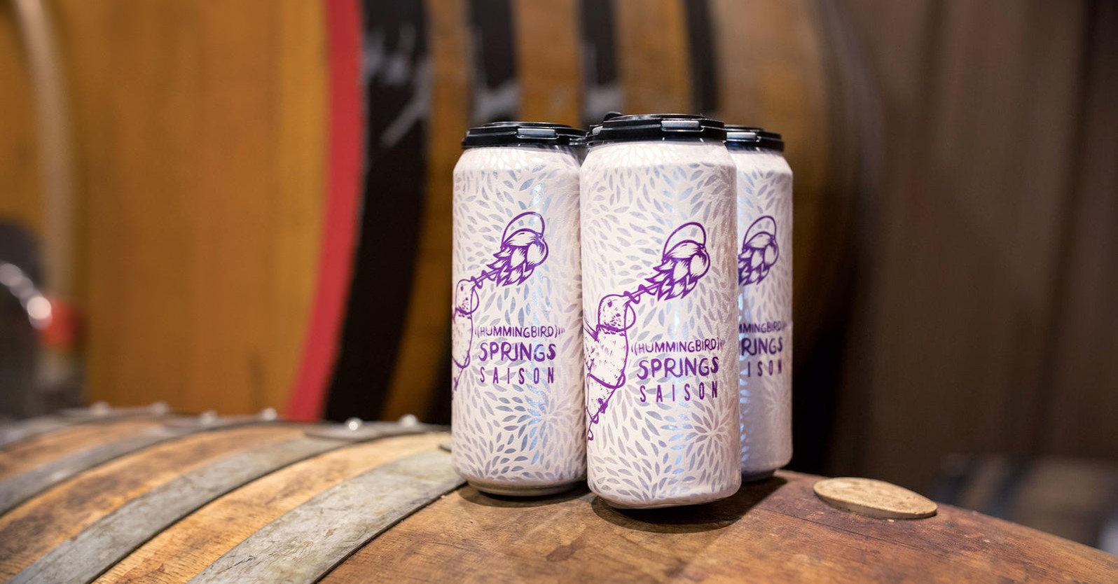 A four-pack of cans from Arizona Wilderness Brewing Co rests on a barrel. The cans have artwork of a hummingbird drinking from a flower and the label states "Hummingbird Springs Saison." 