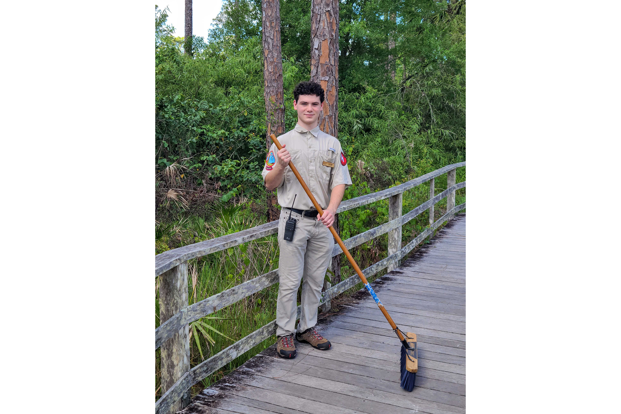 A young man wearing a uniform holds a broom on a boardwalk in nature.