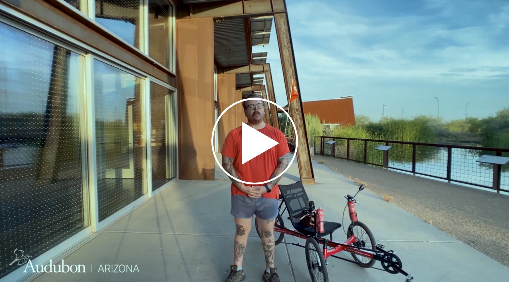 Corey Lycoloplus, a white man with tattoos, stands on a patio under the Rio Salado Audubon Center, with bird-friendly windows to the left and a pond to the right.