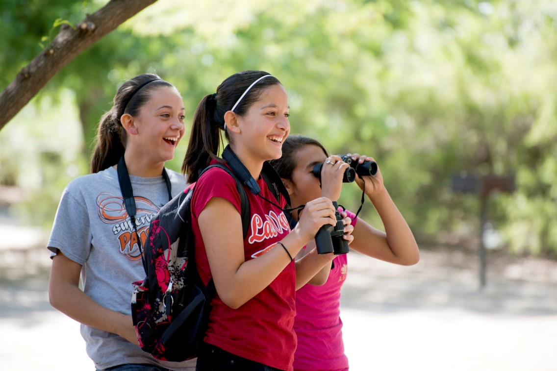 A group of three girls smiles while looking at something with binoculars.