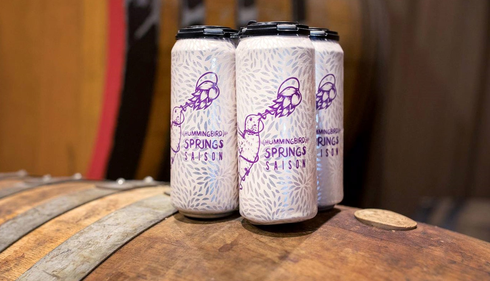 A four-pack of Hummingbird Springs Saison cans sits on top of a barrel.