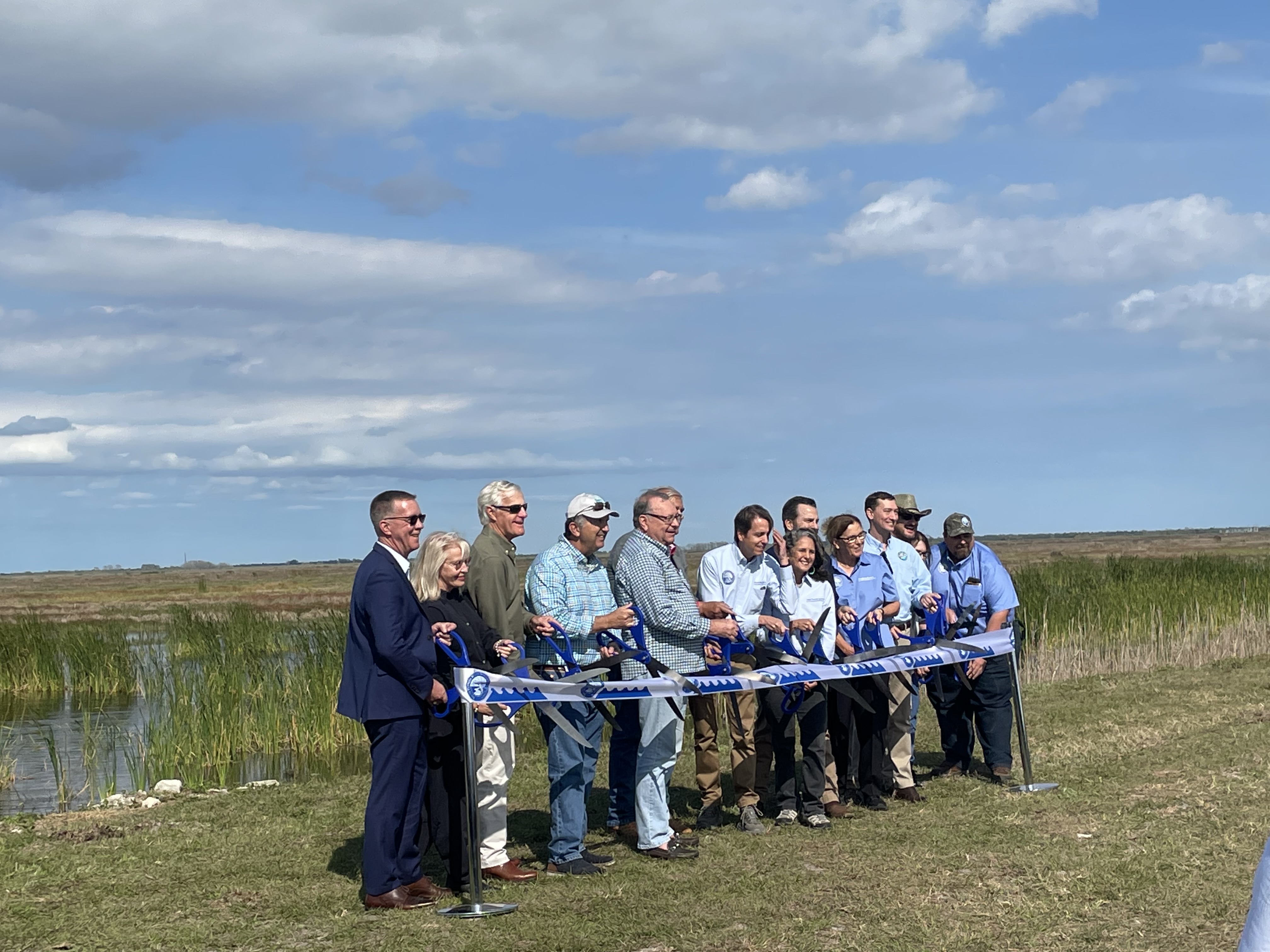 A group of people cutting a ribbon with giant scissors, with a marsh in the background.
