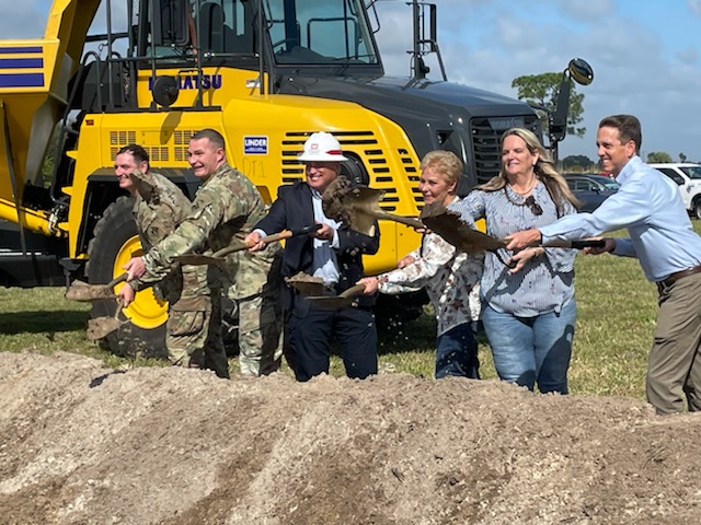 Groundbreaking event. People shovel dirt with a large tractor in the background.