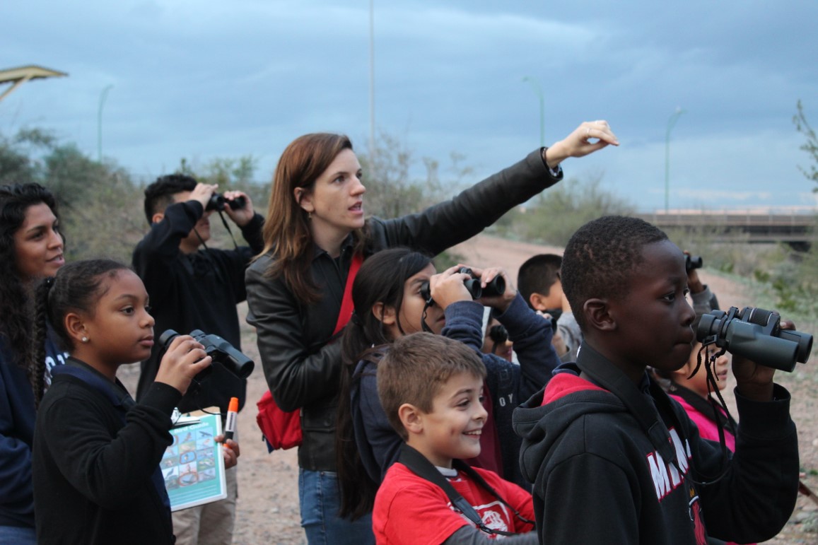 Audubon's teacher/naturalist points to a bird off-screen while children look with binoculars and smile.