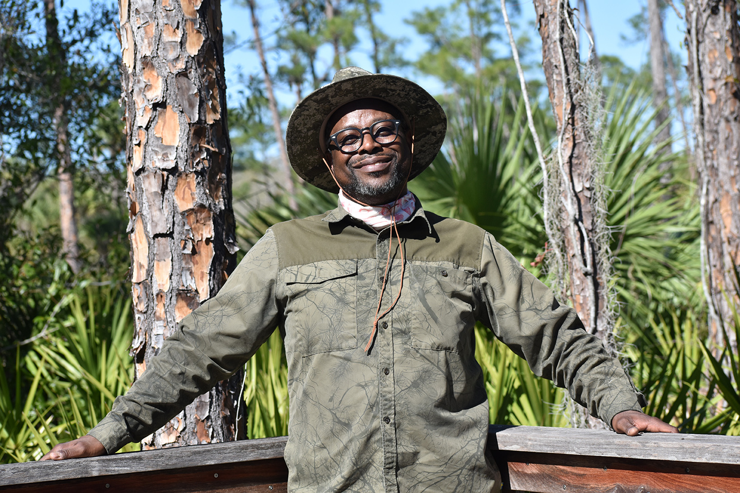 A smiling man wearing camouflage clothes standing in front of a forest.