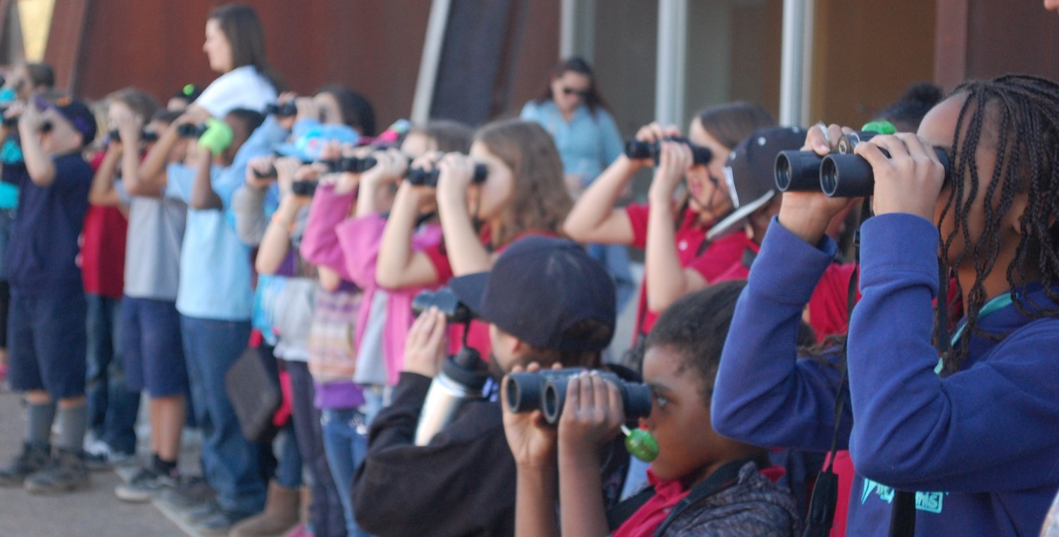 A line of children look out to the sky with binoculars.