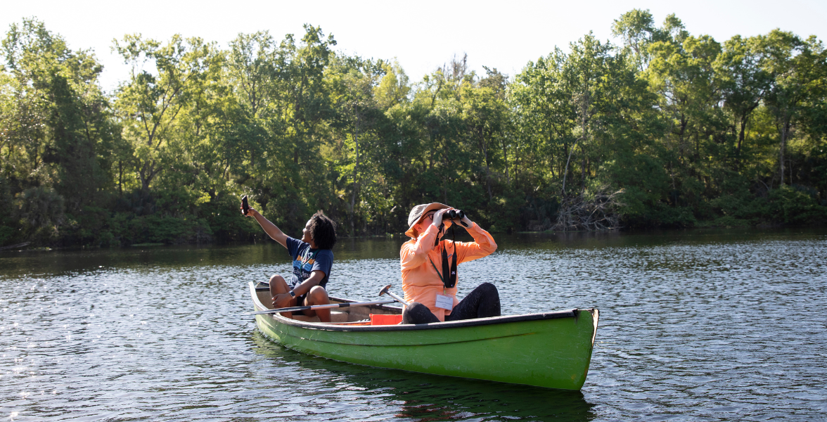 Two people in a green canoe floating on a lake. The person at the front of the canoe is holding binoculars up to their eyes. The person in the back of the canoe is using their phone to take a photo.