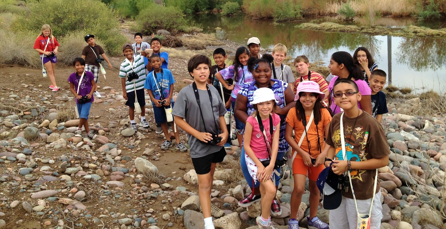 A group of smiling children wave to the camera in front of a river.