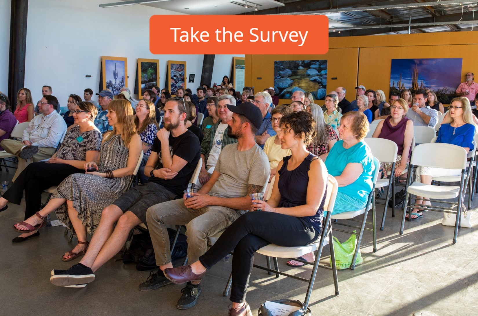 An audience sits in the nature center looking ahead at a speaker off screen. Text reads: "Take the Survey"