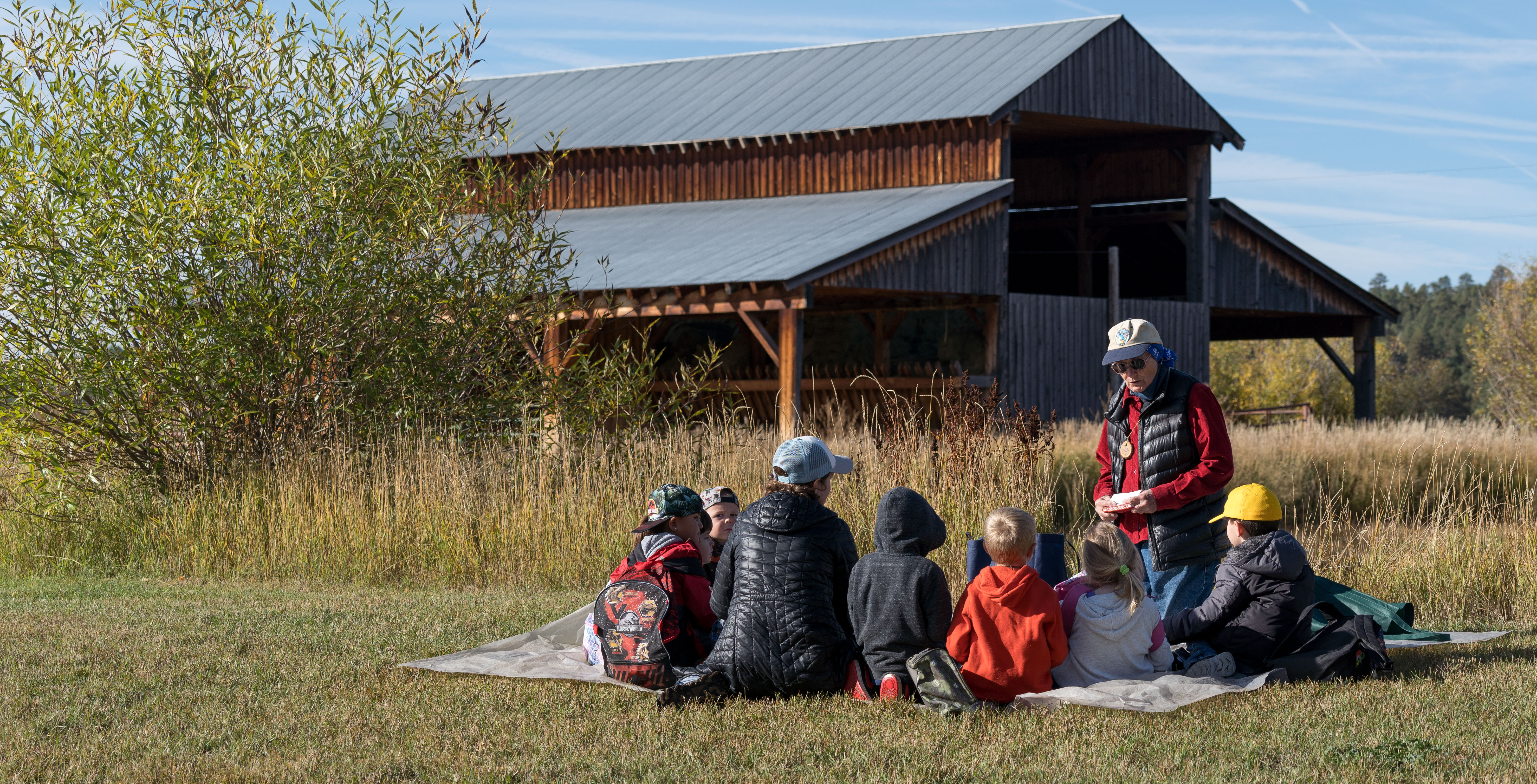 An adult educator is surrounded by young children on a blanket in a grassy field in autumn. A rustic building is in the background.
