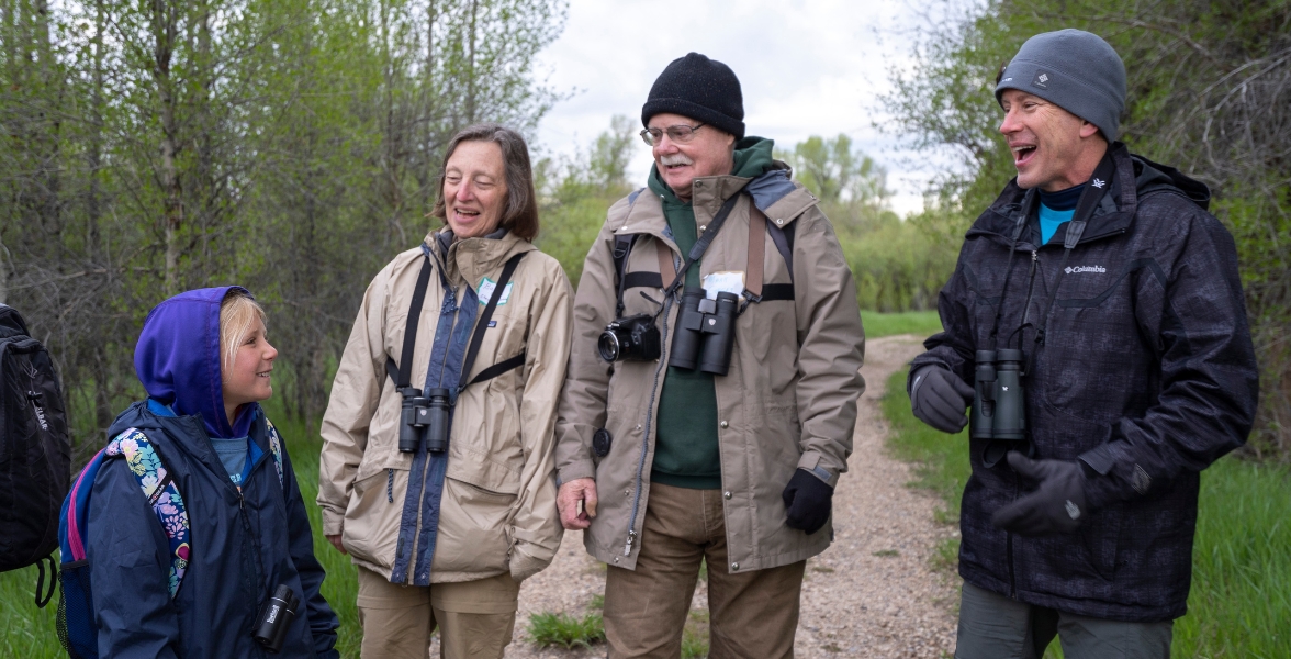 Three adults and a child, all dressed for cold weather, walk down a trail with binoculars. They are all smiling.