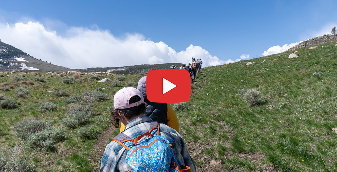 A line of adults hike up a trail. A play button is on the image.