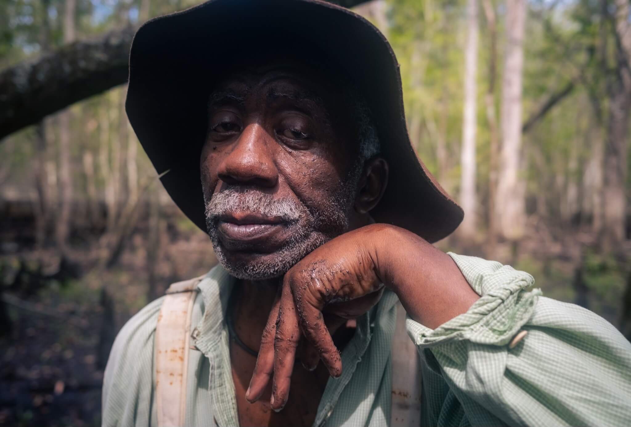 A man in a wide brimmed hat is in a swamp. He is resting his head on his hand. 