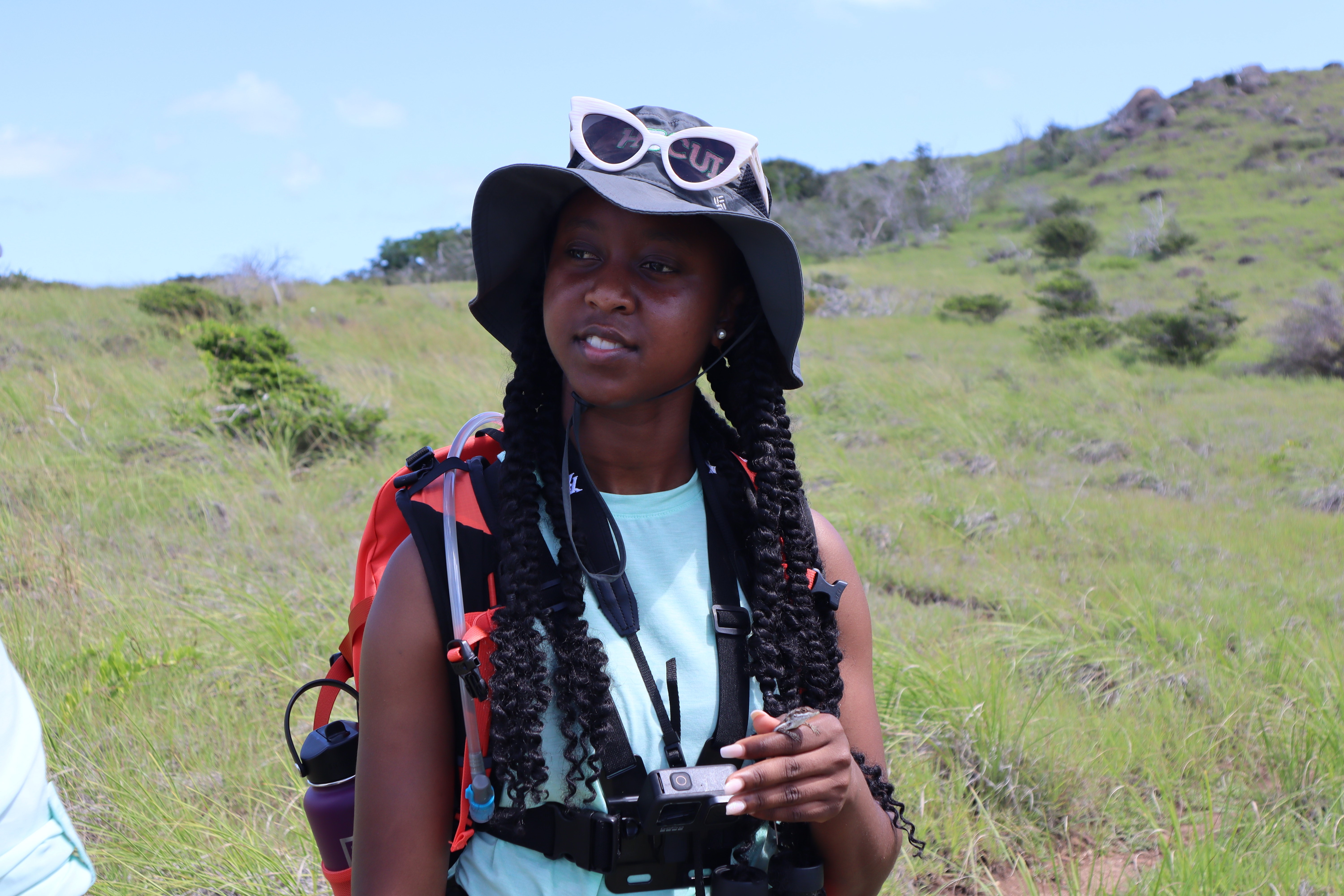 A young woman in field gear walking on a hill carrying a small reptile.